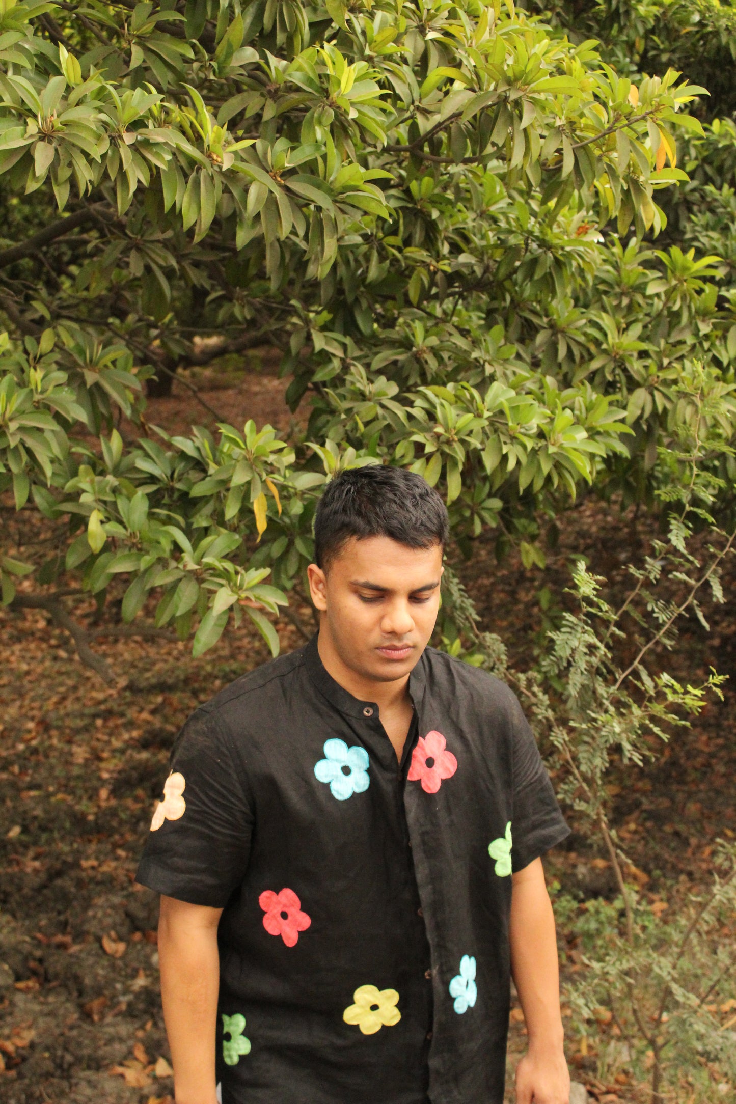 A young man in a black linen shirt with colorful flowers coming out of a grove of trees 