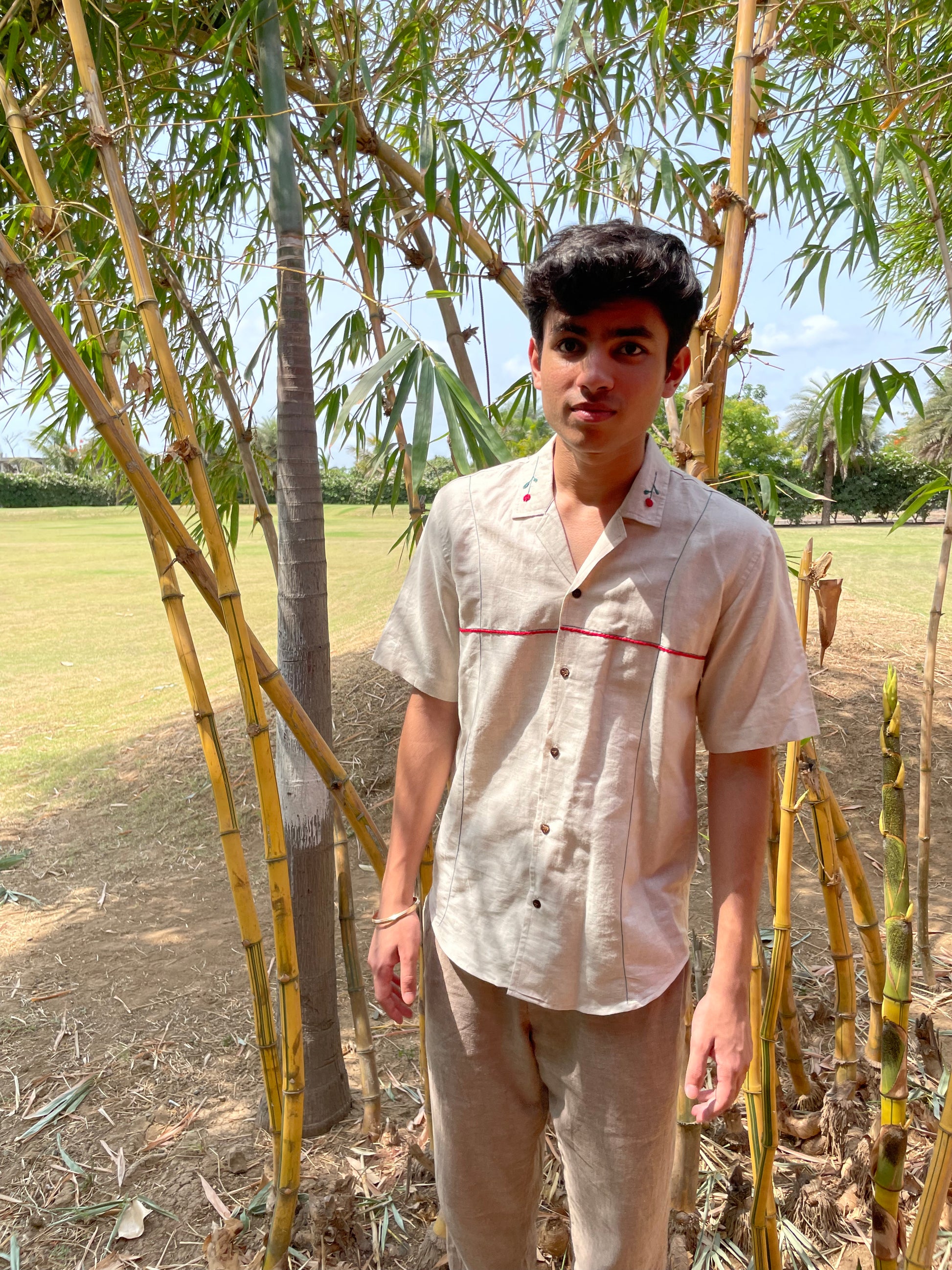 a young man in a beige linen shirt with hand-embroidered roses on the collar is standing in a green field