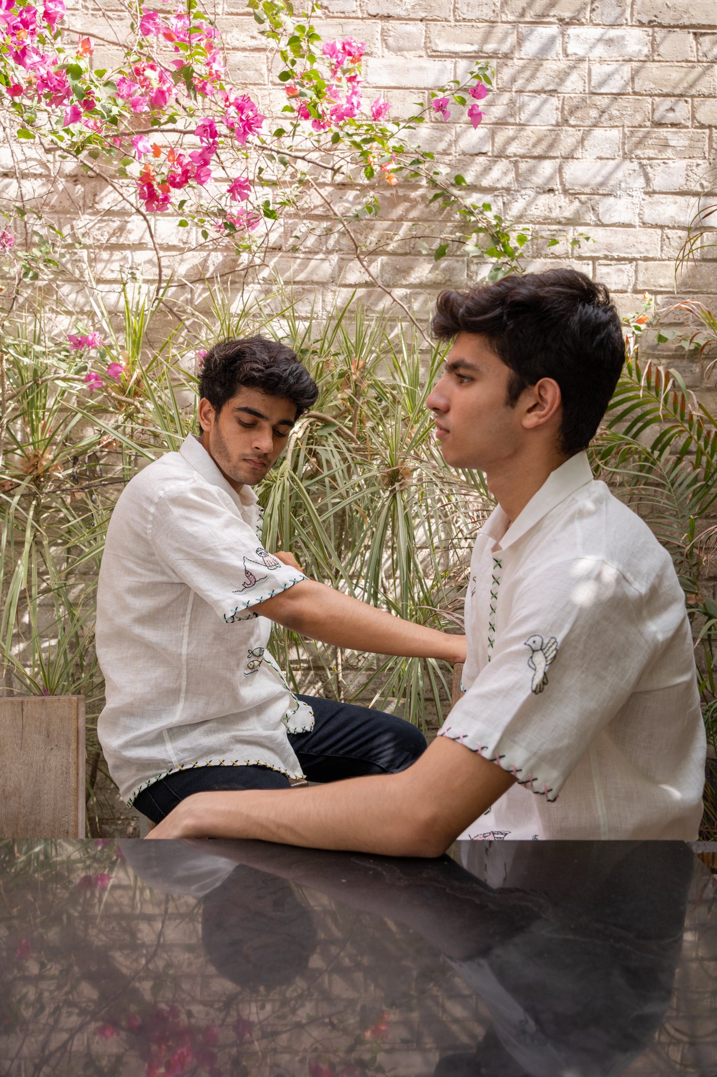 Side profile of two boys wearing the same white linen half-sleeve shirt with multiple motifs like elephants, birds, huts embroidered all over with the focus on sleeve details