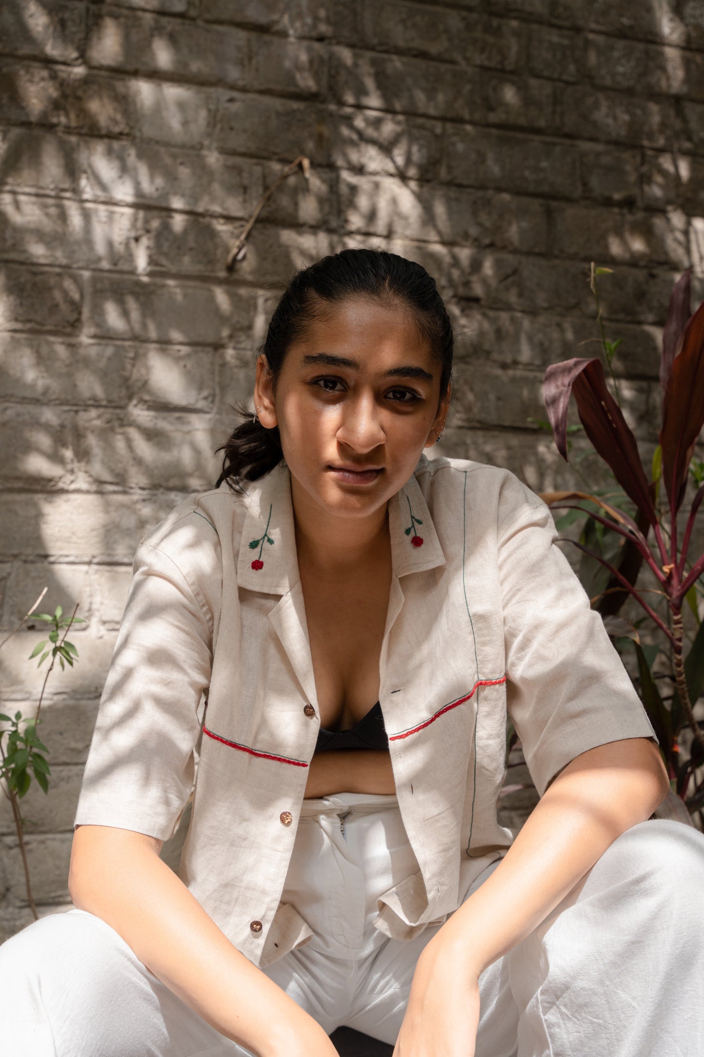 a girl in a beige linen shirt with hand-embroidered roses on the collar is sitting on a chair against an off-white wall background