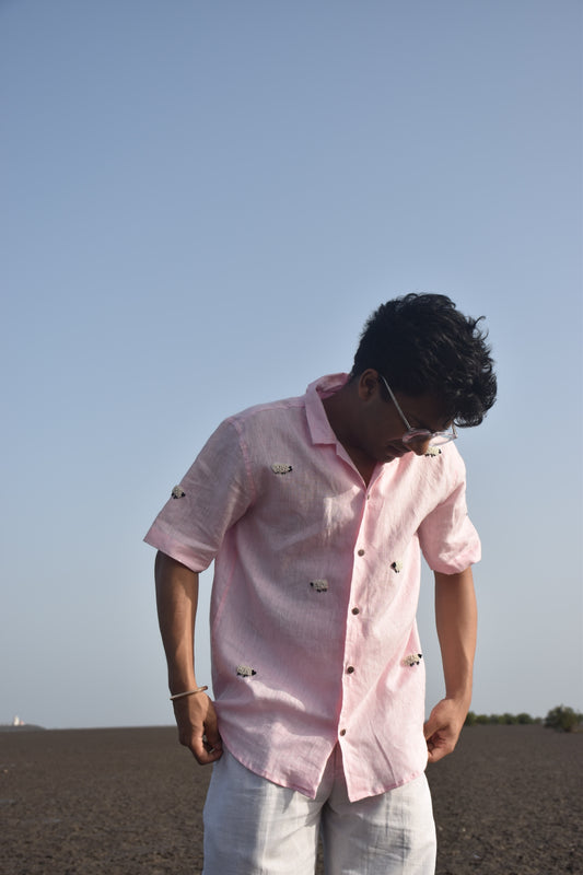 A young man in a pink linen shirt with sheep embroidered throughout is standing on a beach