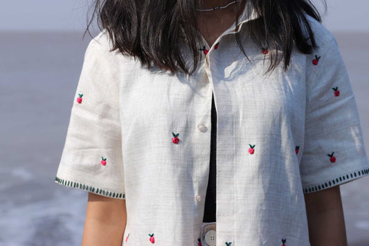 Front shot of a girl in a hand-embroidered off-white linen shirt with small flower buds throughout standing against the backdrop of the sea