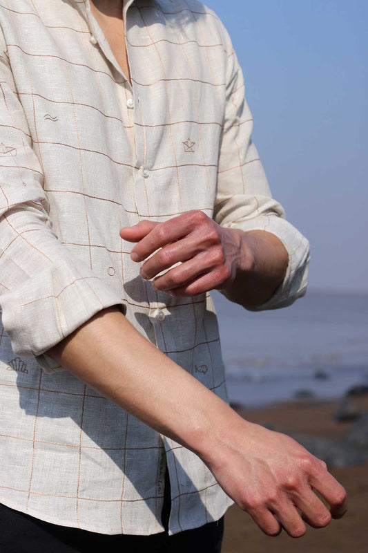 Side view of a young man in a beige linen full sleeved shirt with small embroidered checks and beach elemets like boats, fish shells interspersed throughout