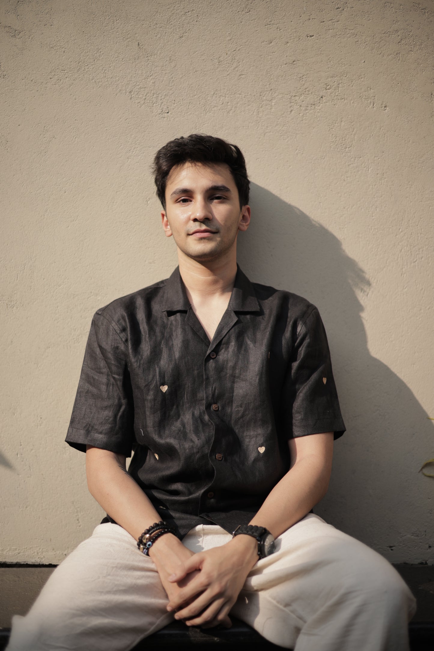 A young man in a black linen shirt with tiny embroidered hearts sitting against a white wall