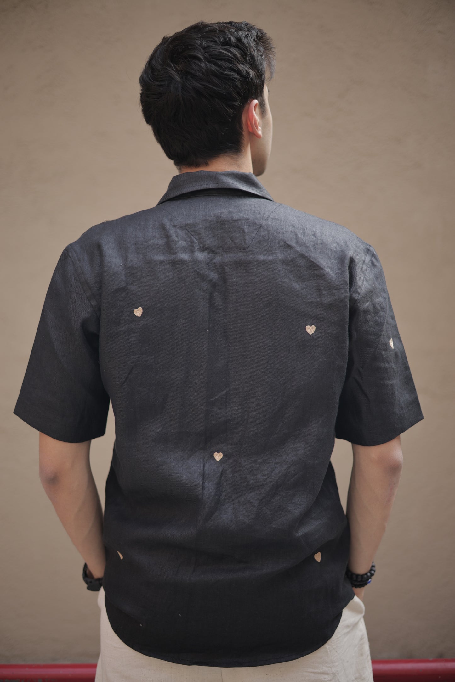 Back of a young man in a black linen shirt with tiny embroidered hearts sitting against a white wall