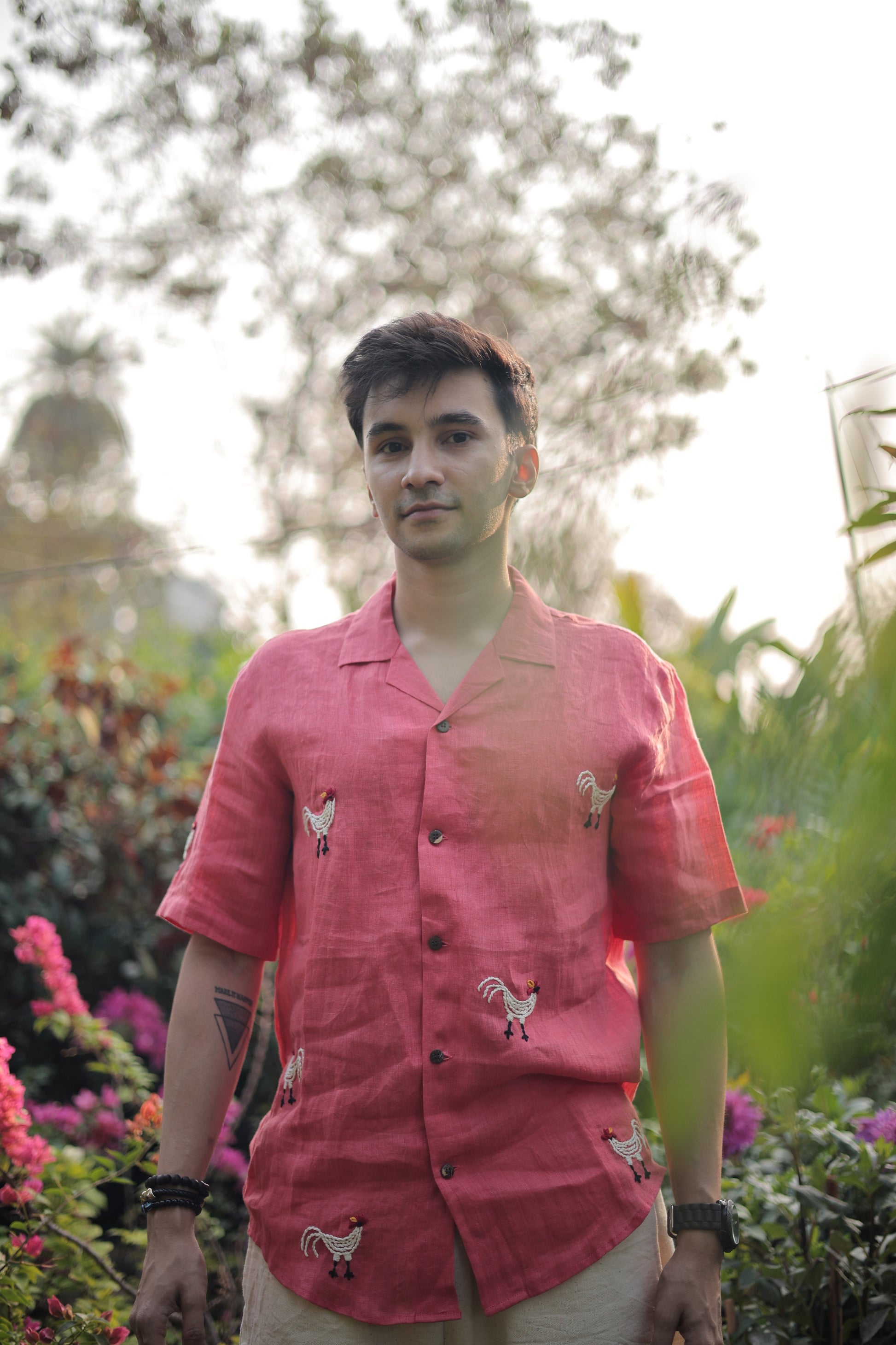 Karan in a beautiful red linen shirt with hand-embroidered roosters is standing in the middle of plants and trees