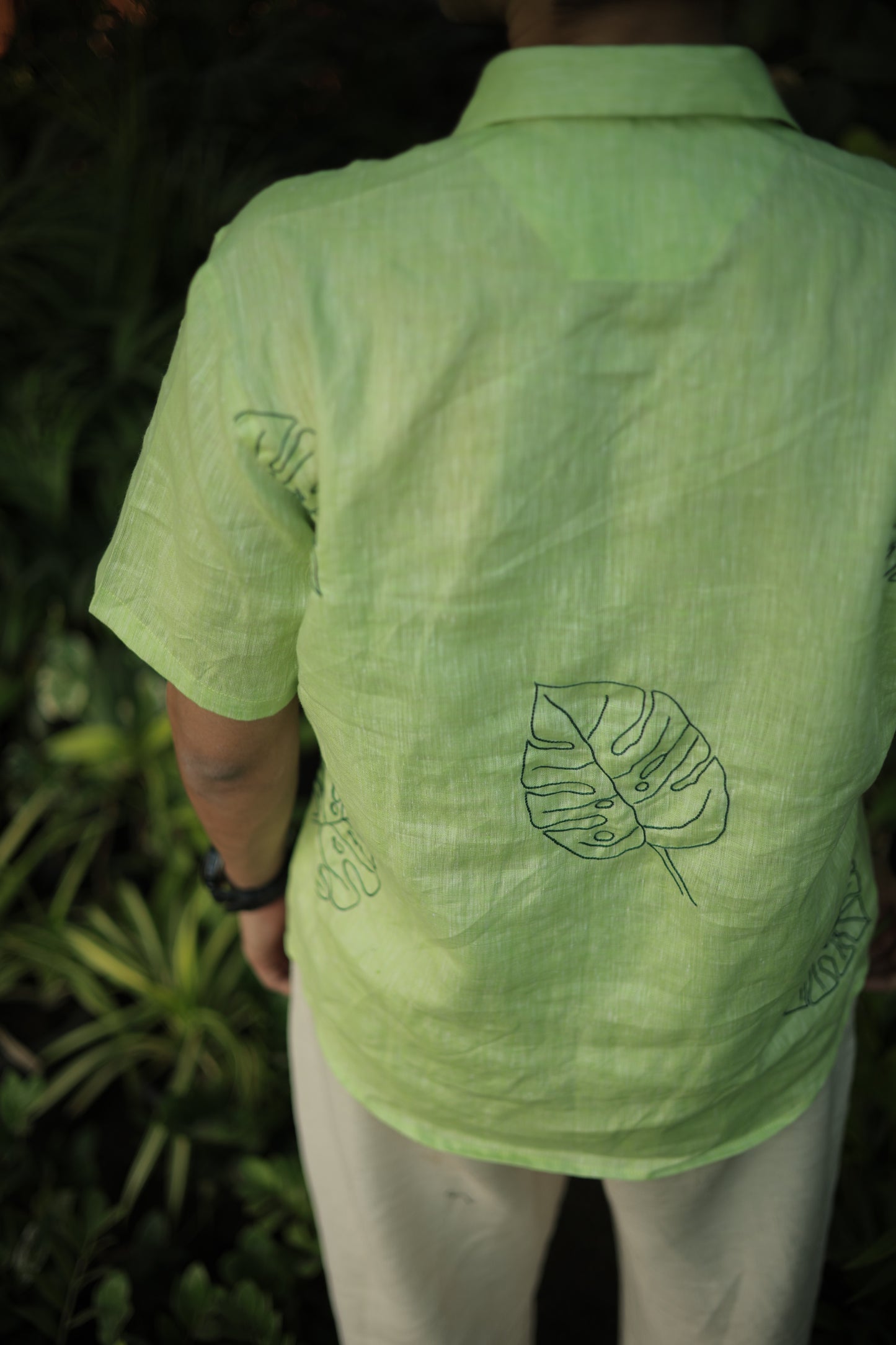 Back detail of a green linen shirt with hand embroidered leaves sitting among different plants