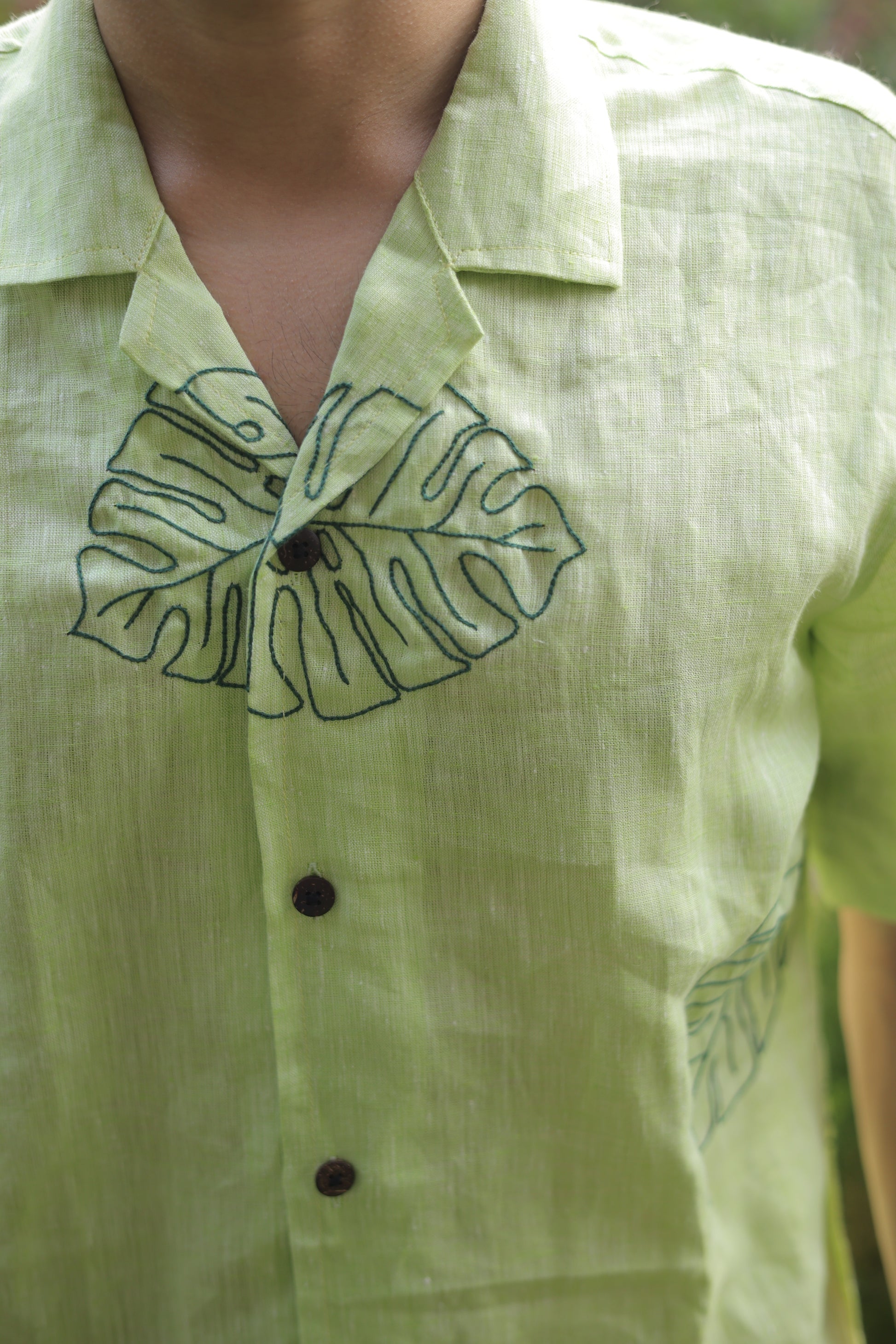 Detail of a green linen shirt with hand embroidered leaves sitting among different plants