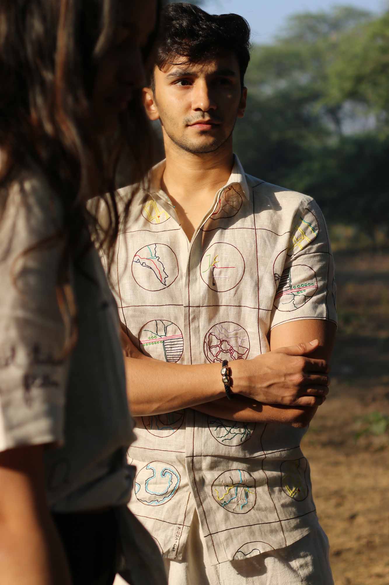 A young man in a beige half-sleeved linen shirt with Hand-embroidered circular maps of cities standing in a field