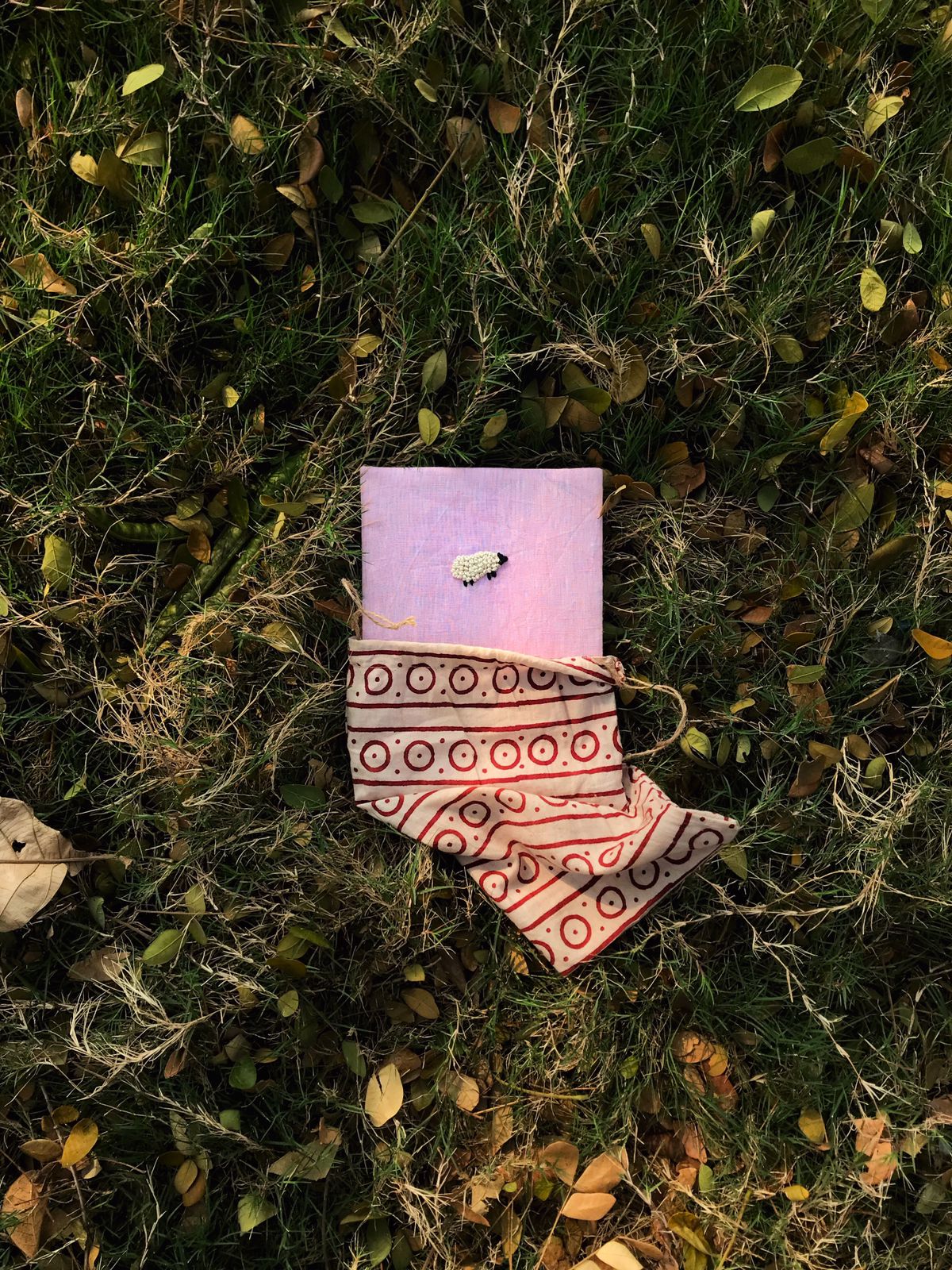 A pink cover diary made with pink linen scraps with a white sheep embroidered on the cover lying amidst green grass