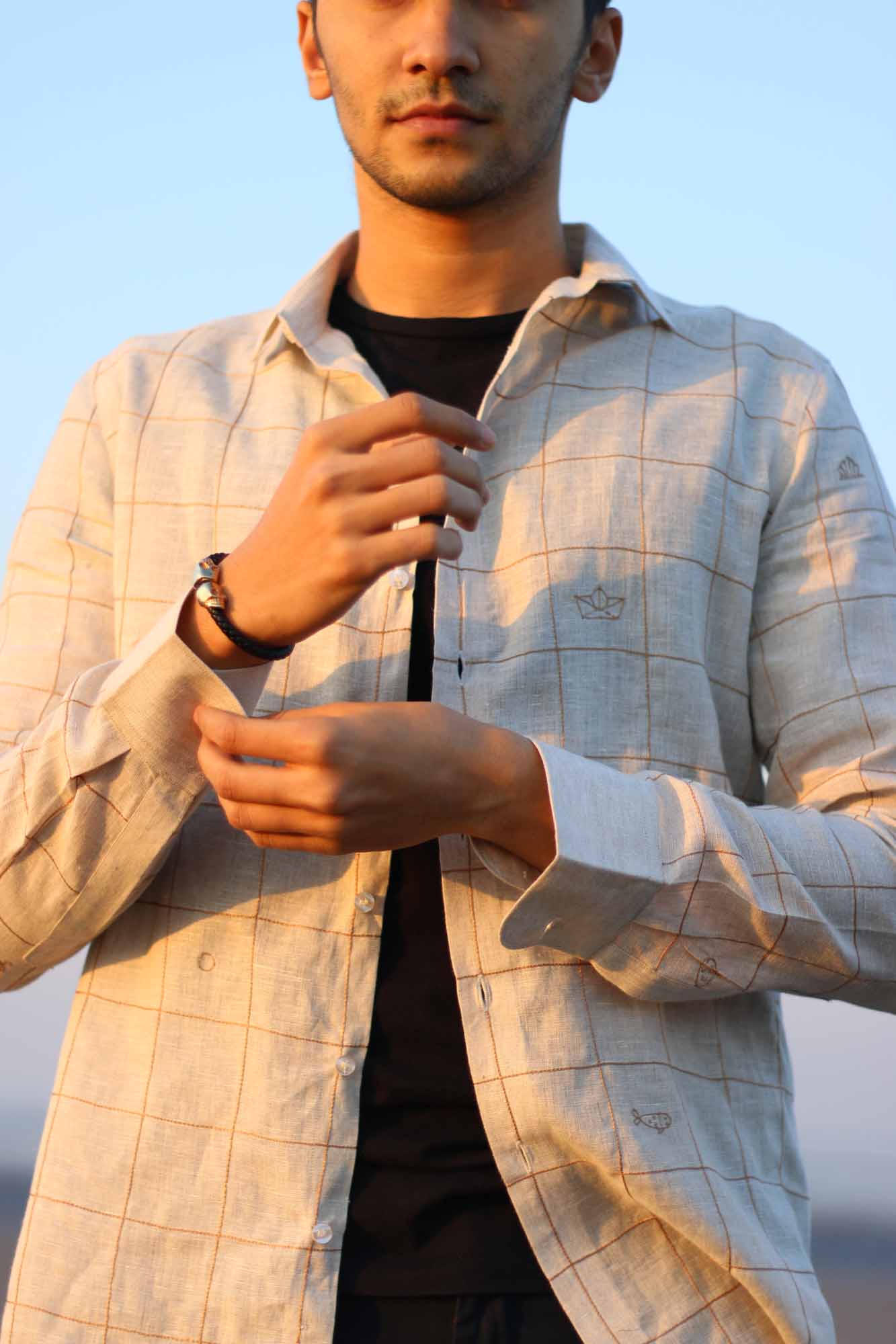 A young man in a beige linen shirt with small embroidered checks and beach elemets like boats, fish shells interspersed throughout